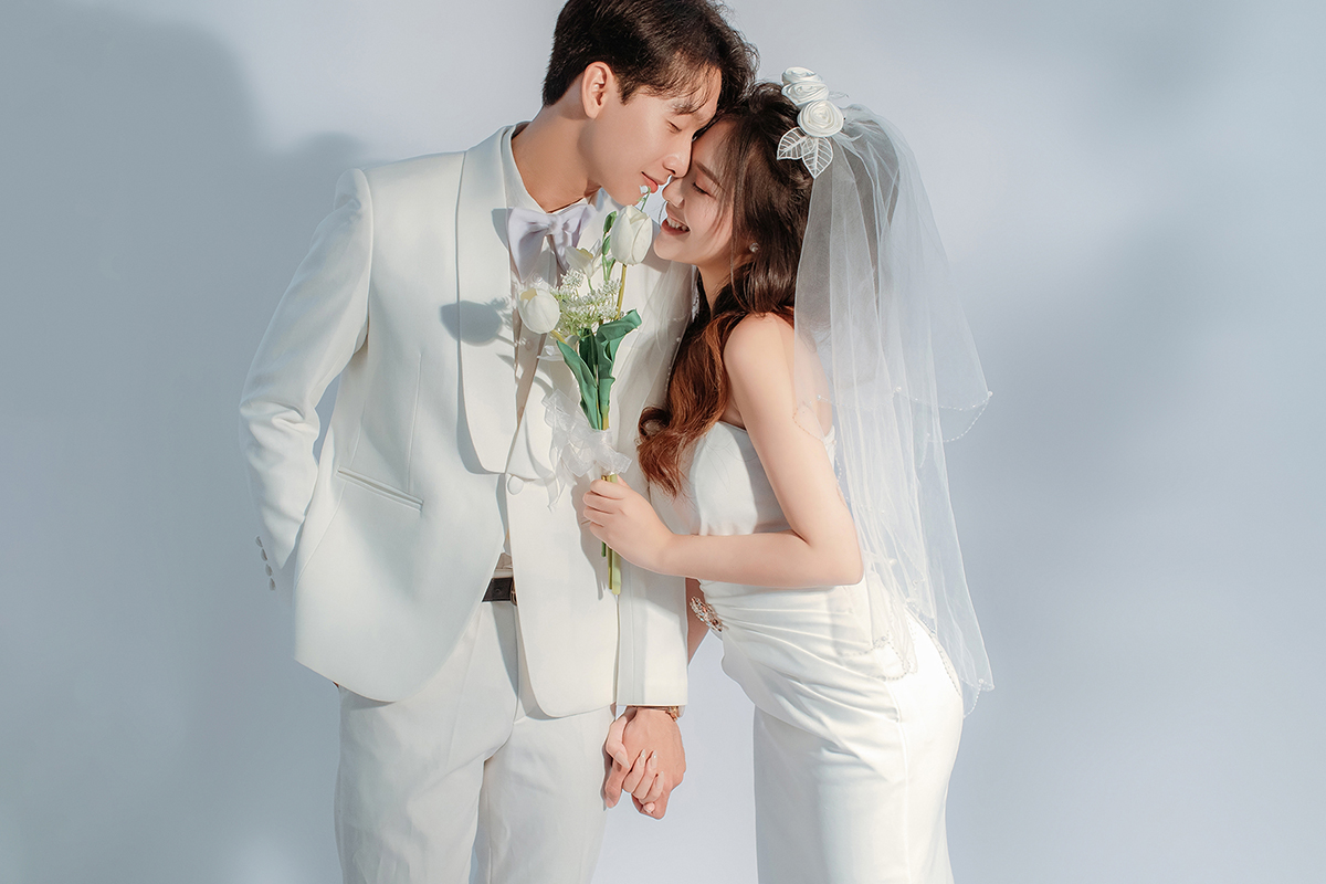 Ảnh cưới phong cách tối giản trên phông trắng trong studio, cô dâu cầm hoa và chú rể vest trắng